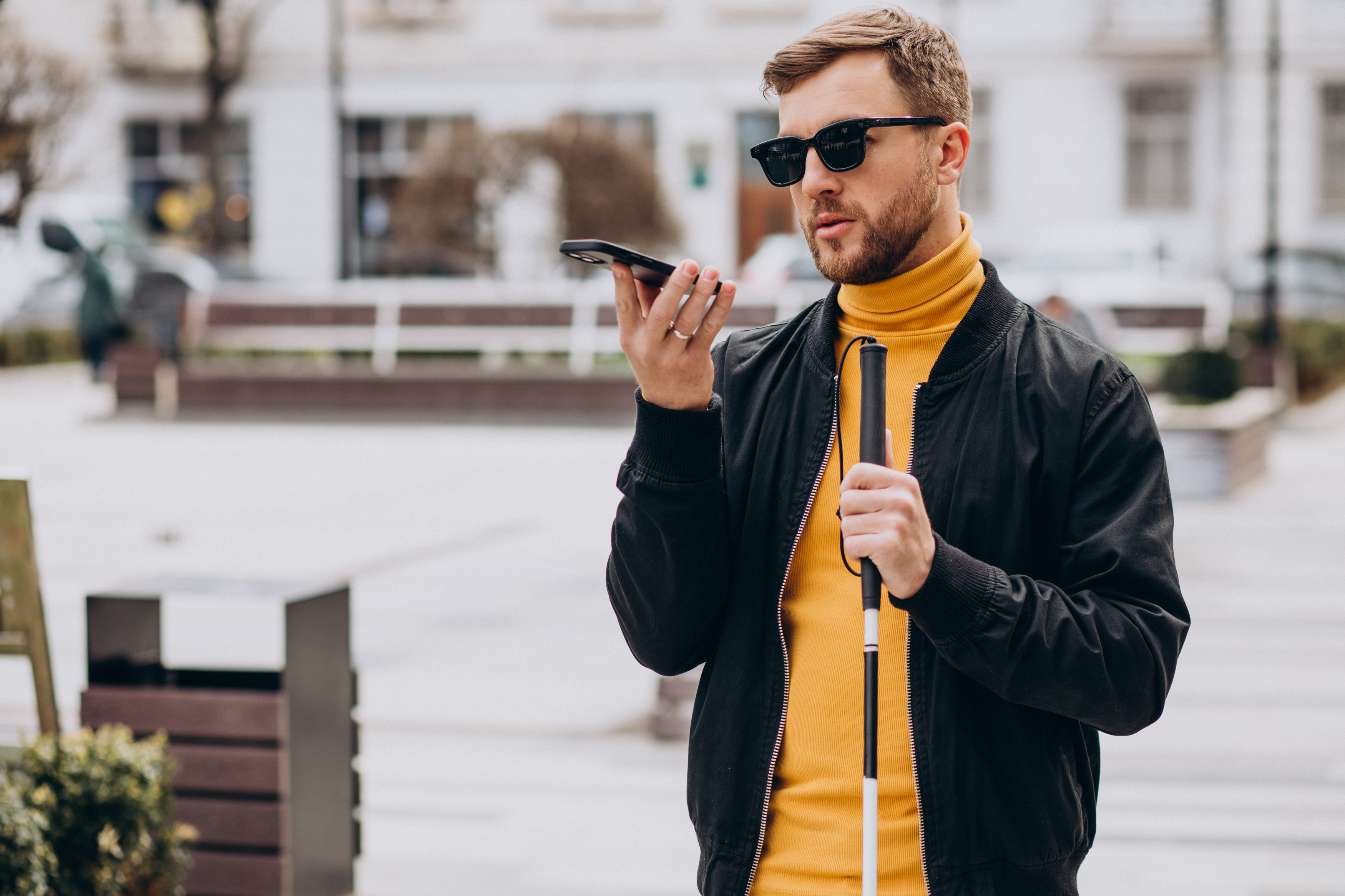 A man speaking into a cellphone. The man is wearing a jacket, turtleneck and sunglasses and carrying a cane.
